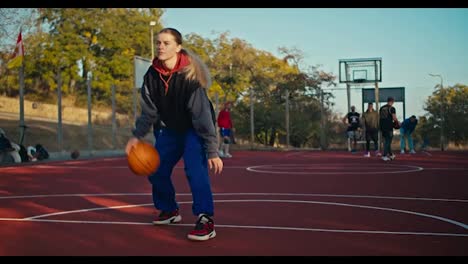 A-blonde-girl-in-a-sports-uniform-hits-an-orange-ball-from-the-floor-during-her-training-on-a-red-street-court-in-the-summer-morning.-Blonde-girl-in-sportswear-in-the-morning-for-basketball-training-on-the-street-court