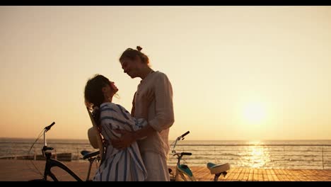 Happy-couple-guy-and-girl-hugging-each-other-on-the-beach-near-the-sea-during-sunrise-in-summer.-A-guy-in-light-clothes-and-a-girl-in-a-white-and-blue-shirt-arrived-at-the-beach-on-bicycles