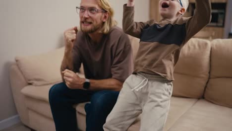 Happy-little-albino-boy-with-white-hair-wearing-blue-glasses-jumps-up-and-throws-a-soccer-ball-while-watching-a-football-match-with-his-blond-father-with-a-beard-and-glasses-at-home-on-the-sofa