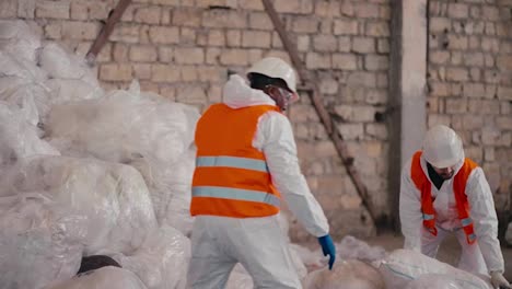 Un-Hombre-De-Piel-Negra-Con-Un-Uniforme-De-Protección-Blanco,-Un-Chaleco-Naranja-Y-Un-Casco-Blanco-Apila-Bolsas-De-Celofán-Y-Polietileno-En-Una-Gran-Pila-Junto-Con-Su-Colega-En-Una-Gran-Planta-De-Procesamiento-Y-Clasificación-De-Residuos.