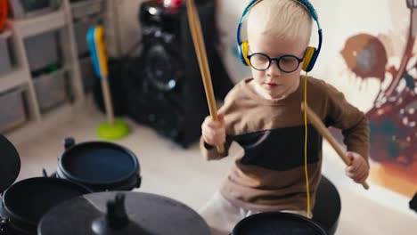 Close-up-shot-from-above-of-a-small-albino-boy-with-white-hair-wearing-blue-glasses-playing-an-electronic-drum-kit-using-special-wooden-sticks-at-home-in-his-room