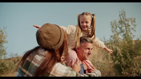 A-little-blonde-girl-has-fun-when-she-sits-on-the-shoulders-of-her-dad,-the-brunette-man-with-a-little-gray-hair-in-a-checkered-shirt,-and-talks-to-her-mom,-the-brunette-girl-in-a-green-checkered-shirt,-during-her-picnic