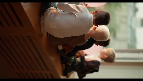 Vertical-video-of-preschool-children-sitting-on-the-windowsill-and-looking-out-the-large-window-at-the-city.-During-a-break-between-classes,-children-look-out-the-window-and-sit-on-the-windowsill