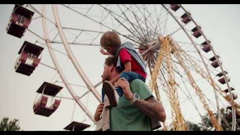 Un-Niño-Rubio-De-Ojos-Azules-Con-Una-Camiseta-Roja-Se-Sienta-Sobre-Los-Hombros-De-Su-Padre-De-Pelo-Rizado-Con-Una-Camiseta-Verde-En-Un-Parque-De-Atracciones-Con-Una-Noria-De-Fondo.