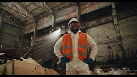 A-confident-and-happy-man-with-Black-skin-in-a-white-helmet-and-a-special-protective-uniform-stands-near-a-large-pile-of-garbage-and-a-conveyor-at-a-huge-waste-recycling-plant