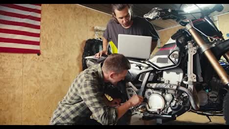 Two-mechanic-guys-are-disassembling-and-inspecting-a-motorcycle.-A-guy-in-a-gray-T-shirt-looks-at-a-laptop-screen-in-a-workshop-with-tools.-Service-center-for-equipment