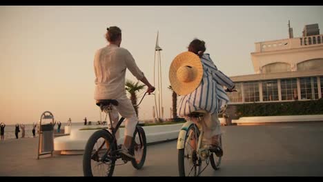 Rear-view-of-a-happy-guy-in-white-clothes-on-a-Black-bicycle-and-a-girl-with-a-Straw-hat-on-a-bicycle-ride-along-the-beach-near-the-sea-at-sunrise-in-summer.-Active-leisure-and-date-on-the-go-on-bicycles-near-the-sea-on-the-beach