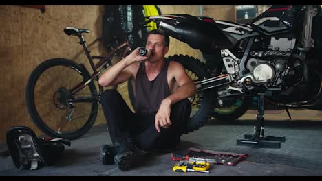 A-male-mechanic-with-a-short-haircut-in-a-gray-T-shirt-drinks-a-drink-from-a-dark-glass-bottle-while-sitting-near-his-motorcycle-in-his-workshop.-Rest-during-a-break-during-motorcycle-repair-and-maintenance