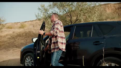 A-happy-man-with-gray-hair-in-a-plaid-shirt-gets-out-of-his-black-one-in-the-car-and,-together-with-his-girlfriend-and-daughter,-takes-out-things-for-a-picnic-in-a-steppe-area-with-dry-grass-in-summer