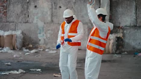 Un-Hombre-De-Piel-Negra-Con-Un-Uniforme-De-Protección-Blanco-Y-Un-Chaleco-Naranja-Junto-Con-Su-Colega-Un-Hombre-Con-Barba-Con-Un-Casco-De-Protección-Blanco-Baila-En-El-Gran-Salón-De-Una-Planta-De-Procesamiento-Y-Clasificación-De-Residuos.