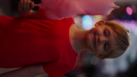 Vertical-video-portrait-of-a-happy-little-blond-boy-with-blue-hair-in-a-red-T-shirt-who-looks-appetizingly-at-pink-cotton-candy-and-bites-it-in-the-amusement-park