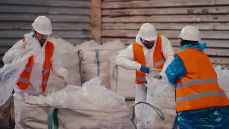 A-team-of-three-workers-in-a-white-uniform-and-an-orange-vest-stack-and-press-polyethylene-and-cellophane-while-working-at-a-large-waste-recycling-plant