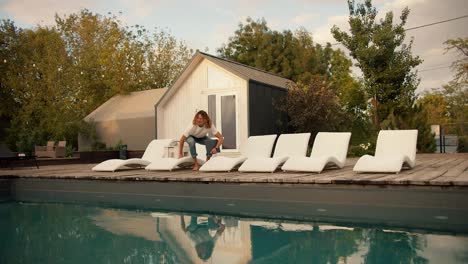 A-curly-haired-guy-in-glasses-falls-on-a-white-sun-lounger-near-the-sunbeds-by-the-pool-and-opens-a-laptop.-Rest-in-the-country-house