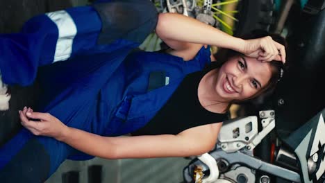 Vertical-video:-Vertical-video:-Portrait-of-a-brunette-mechanic-girl-in-a-black-top-and-blue-overalls-looking-at-the-camera-and-smiling.-Girl-mechanic-sits-on-the-floor-and-leans-on-a-bike