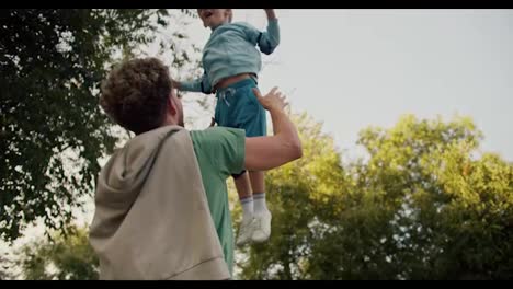 A-father-with-curly-hair-in-a-Green-T-shirt-throws-his-little-blond-son-in-a-Blue-Jacket-up-in-the-park.-A-fun-attraction-for-a-little-boy.-Happiness-in-the-family-between-father-and-son