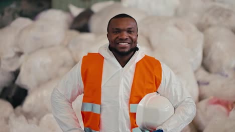 Portrait-of-a-Happy-man-with-Black-skin-in-a-white-protective-uniform-who-holds-in-his-hands-a-protective-white-helmet-and-an-orange-vest-on-it.-He-stands-near-large-bags-of-cellophane-and-polyethylene-at-a-waste-recycling-plant