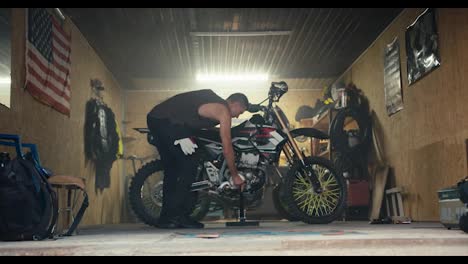 A-male-mechanic-with-a-short-haircut-and-a-gray-T-shirt-puts-his-motorcycle-on-a-jack-in-his-garage-workshop-against-of-the-US-flag.-Start-of-the-working-day-and-start-of-motorcycle-maintenance-in-the-workshop