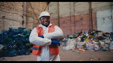 Retrato-De-Un-Hombre-De-Mediana-Edad-Feliz,-De-Piel-Negra,-Con-Un-Uniforme-Blanco-Y-Un-Chaleco-Naranja,-Que-Se-Encuentra-Frente-A-Una-Enorme-Pila-De-Basura-Plástica-Y-Desechos-En-Una-Planta-De-Reciclaje-De-Desechos.