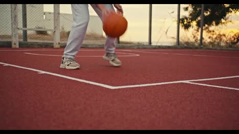 Ein-Mädchen-In-Weißen-Hosen-Lässt-Einen-Orangefarbenen-Basketball-Auf-Einem-Roten-Basketballplatz,-Der-Mit-Gittern-Umzäunt-Ist,-Frühmorgens-Bei-Sonnenaufgang-Vom-Boden-Aufprallen.