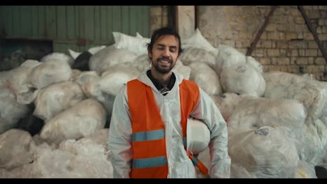 Portrait-of-a-happy-and-confident-brunette-man-with-a-beard-in-a-white-protective-helmet-and-an-orange-vest-posing-against-the-backdrop-of-huge-piles-of-recycled-and-sorted-cellophane-at-a-waste-processing-and-sorting-plant