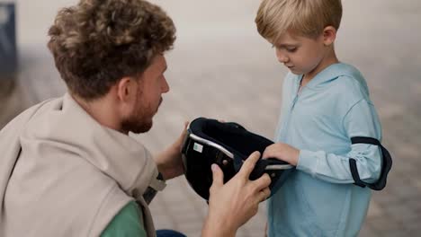 Ein-Vater-Mit-Lockigem-Haar-In-Einem-Grünen-T-Shirt-Hilft-Seinem-Kleinen-Sohn,-Zum-Schutz-Einen-Schwarzen-Helm-Aufzusetzen,-Während-Er-Im-Sommer-In-Einem-Skatepark-Skateboard-Fährt