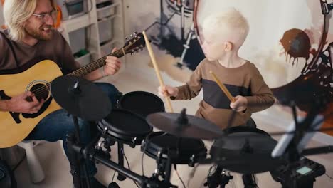 Ein-Glücklicher-Albino-Junge-Mit-Weißem-Haar-In-Einem-Braunen-Pullover-Spielt-Auf-Einem-Elektronischen-Schlagzeug-Mit-Seinem-Blonden-Vater-Mit-Brille-Und-Bart,-Der-An-Seinem-Freien-Tag-Mit-Seinem-Kleinen-Sohn-Im-Zimmer-Akustikgitarre-Spielt