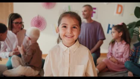 Retrato:-Una-Niña-Feliz-Con-Ojos-Marrones-Y-Una-Camisa-Blanca-Mira-A-La-Cámara-Y-Sonríe-En-Su-Primera-Lección-En-El-Club-Preescolar.-La-Niña-Se-Sienta-En-Su-Primera-Lección-En-El-Club-De-Preparación-Escolar.