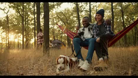 A-happy-couple,-a-man-with-Black-skin-and-a-girl-in-a-black-hat-in-a-plaid-shirt,-are-sitting-on-a-Red-hammock,-next-to-them-is-their-white-brown-dog,-and-behind-them-is-the-rest-of-the-group-of-people-who-are-going-on-a-hike-and-in-a-tent-in-a-green-Sunny-forest-during-the-evening