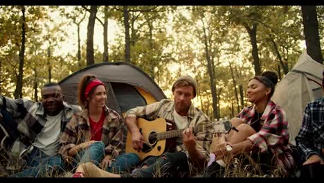 Un-Hombre-Rubio-Toca-La-Guitarra-Mientras-El-Resto-De-Excursionistas,-Durante-Una-Parada,-Cantan-Con-él-Y-Escuchan-Su-Agradable-Música-En-La-Guitarra-Con-El-Telón-De-Fondo-De-Tiendas-De-Campaña-En-Un-Soleado-Bosque-Verde-De-Verano.