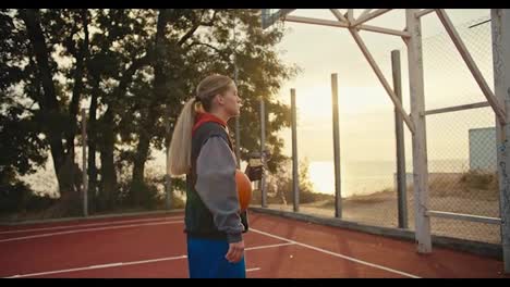 Close-up-a-blonde-girl-drinks-water-from-a-sports-bottle-and-holds-an-orange-basketball-in-her-hands-during-her-workout-in-the-morning-at-Sunrise-in-the-summer