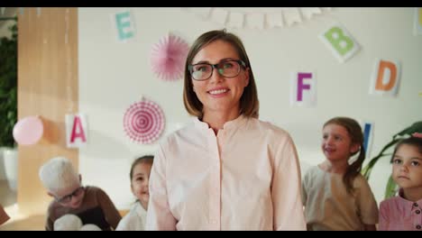 Retrato-De-Una-Profesora-Feliz-Con-Un-Peinado-Bob,-Gafas-Y-Una-Camisa-Blanca-Que-Mira-A-La-Cámara-Y-Sonríe-Ampliamente-En-El-Contexto-De-Un-Grupo-De-Niños-En-Un-Club-De-Preparación-Escolar.