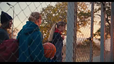 A-happy-blonde-girl-together-with-her-friends-with-a-basketball,-enters-a-special-basketball-court-fenced-with-a-grid-for-playing-basketball-early-in-the-morning-at-sunrise