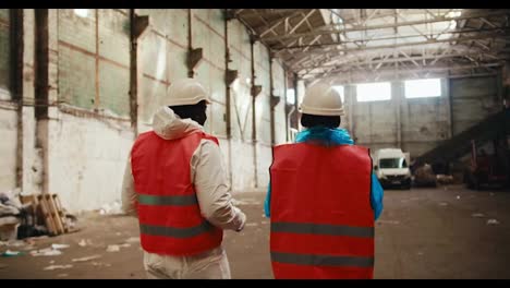 Rear-view-of-a-man-with-Black-skin-in-a-white-special-uniform-and-an-orange-vest-together-with-a-female-employee-walk-through-a-large-waste-processing-plant-and-point-with-his-hand-to-the-side