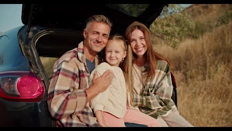 Portrait-of-a-happy-family-who,-during-their-picnic-outside-the-city,-sits-in-the-open-trunk-of-a-black-car-and-poses.-A-happy-brunette-girl,-together-with-her-husband,-the-middle-aged-man-with-gray-hair-in-a-checkered-shirt,-and-their-little-daughter,-the-blonde-girl,-are-sitting-in-the-open-trunk-