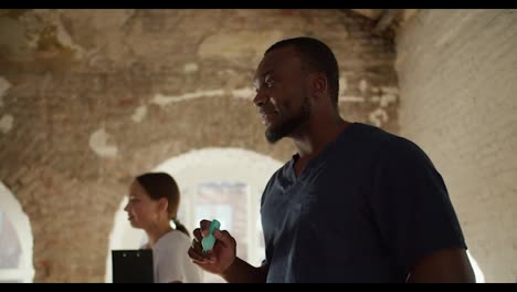 Close-up:-A-Black-male-doctor-in-a-blue-uniform-with-a-green-felt-tip-pen-in-his-hand-gives-a-medical-lecture-and-addresses-his-assistant,-a-female-nurse-in-a-white-uniform,-who-stands-behind-him-in-a-spacious-room-with-white-brick-walls