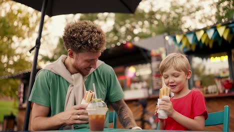 A-father-with-curly-hair-with-stubble-in-a-green-T-shirt-tickles-his-little-blond-son-in-a-red-T-shirt-while-they-eat-hot-dogs-sitting-at-an-outdoor-table-in-a-street-cafe-in-the-park