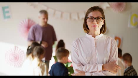 Porträt-Einer-Lehrerin-Mit-Brille-Und-Rosa-Hemd,-Die-Mit-Verschränkten-Armen-Vor-Der-Brust-Lächelt,-Vor-Dem-Hintergrund-Einer-Biologiestunde-Für-Vorschulkinder,-Zusammen-Mit-Einem-Männlichen-Lehrer-In-Einem-Lila-Hemd