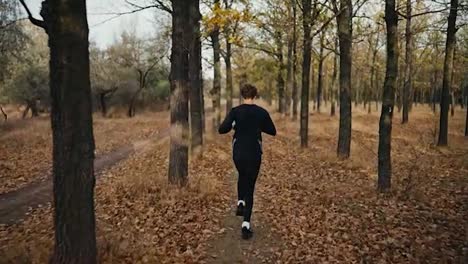 Rear-view-of-a-happy-confident-male-athlete-running-in-a-black-sports-uniform-along-an-earthen-path-in-an-autumn-forest-with-fallen-brown-leaves