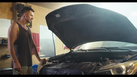 A-man-with-a-short-haircut-in-a-gray-T-shirt-inspects-the-hood-of-his-car-in-his-garage-workshop.-Car-repair-work-and-service