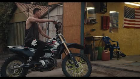 A-male-motorcyclist-with-a-short-haircut-in-a-gray-T-shirt-rides-his-bike-into-his-garage-workshop-which-is-filled-with-various-tools-and-he-puts-his-bike-on-a-jack