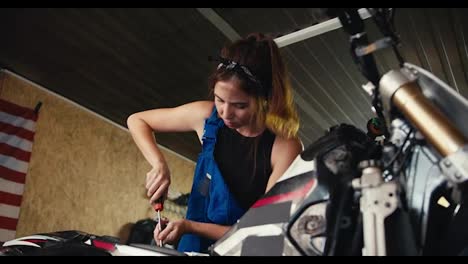 Zoom-in:-a-girl-auto-mechanic-repairs-a-motorcycle-and-shakes-sweat-from-her-forehead-with-her-hand-in-her-workshop.-Tired-of-long-work-in-the-garage-workshop