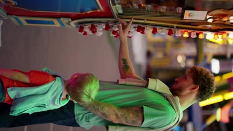 Vertical-video-of-a-happy-dad-with-curly-hair-in-a-green-T-shirt-and-his-little-blond-son-in-a-red-T-shirt-looking-at-the-ticket-office-in-an-amusement-park-and-ordering-tickets-for-themselves