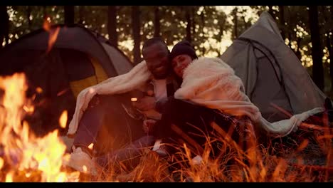 Happy-couple:-a-girl-with-a-bob-hairstyle-examines-the-face-of-a-guy-with-black-skin,-touches-him-and-leans-on-his-shoulder,-they-hug-under-a-white-blanket,-sit-near-a-bright-fire-against-the-backdrop-of-tents-and-a-green-forest-in-autumn