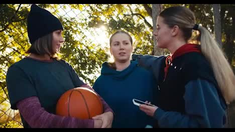 Happy-three-blonde-girls-in-sportswear-communicate-and-hold-an-orange-basketball-ball-on-a-summer-street-playground-near-green-trees-on-a-summer-sunny-day