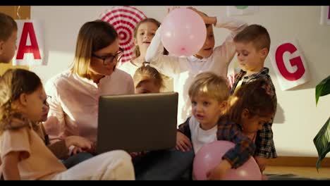 A-female-teacher-with-a-bob-hairstyle,-glasses,-a-fibrous-shirt,-and-a-laptop-are-teaching-a-lesson-for-preschool-children-in-a-special-club-for-preparing-children-for-school