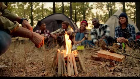 A-small-fire-burns-during-a-halt-near-a-group-of-people-on-a-hike-in-special-hiking-clothes-against-the-backdrop-of-tents-in-a-green-autumn-forest-with-withered-grass.-A-man-sits-near-a-fire-and-warms-his-hands-during-a-stop-on-a-hike