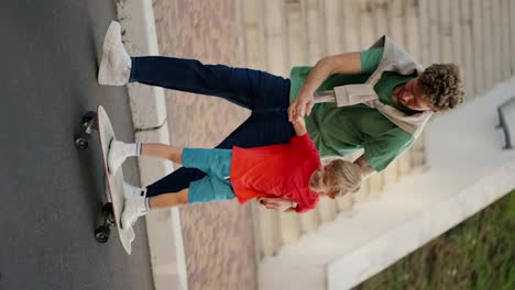 Vertical-video-of-a-dad-with-curly-hair-in-a-Green-T-shirt-and-blue-jeans-helping-his-little-blond-son-in-a-red-T-shirt-and-shorts-ride-a-white-skateboard-along-the-road-in-the-park