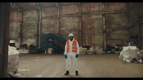 Portrait-of-a-happy-and-confident-man-with-Black-skin-color-in-white-protective-clothing-and-an-orange-vest-who-stands-and-crosses-his-arms-on-his-chest-in-a-huge-waste-recycling-and-sorting-plant