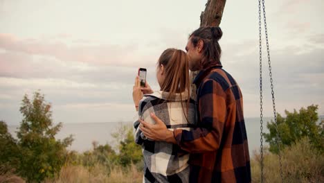 Aufnahme-Von-Hinten:-Ein-Typ-Im-Karierten-Hemd-Umarmt-Und-Streichelt-Seine-Freundin-Im-Karierten-Hemd,-Während-Sie-Mit-Ihrem-Telefon-Ein-Foto-Einer-Meereslandschaft-Macht.-Erholung-Im-Landhaus