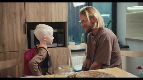 Happy-little-albino-boy-with-white-hair-color-with-a-red-backpack-on-his-back-communicates-with-his-dad,-a-blond-man-with-a-beard-and-glasses-in-the-kitchen-after-breakfast-before-the-start-of-the-school-day-at-school